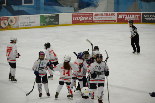 Eine Gruppe junger Menschen, die Eishockey auf einem Eisplatz spielen, alle mit Helmen und Hockeystöcken, mit einer Person auf der rechten Seite und Tafeln mit Text im Hintergrund.