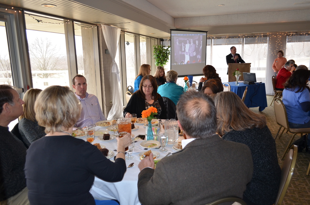 Eine Gruppe von Menschen, die um einen Tisch sitzen, der mit verschiedenen Speisen gefüllt ist, eine Person, die spricht und ein Projektor, der Inhalt anzeigt, eine Pflanze neben dem Projektor und ein Fenster mit Vorhängen, der Himmel und Bäume im Hintergrund.