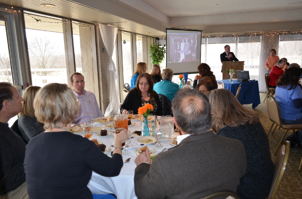 Eine Gruppe von Menschen, die um einen Tisch sitzen, der mit verschiedenen Speisen gefüllt ist, eine Person, die spricht und ein Projektor, der Inhalt anzeigt, eine Pflanze neben dem Projektor und ein Fenster mit Vorhängen, der Himmel und Bäume im Hintergrund.