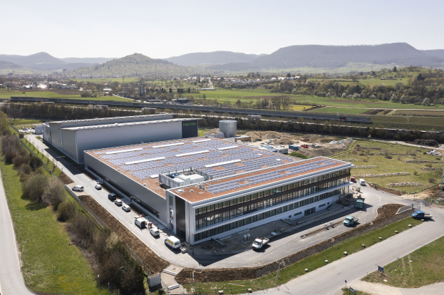 Luftaufnahme eines großen Gebäudes mit Solarpanelen auf dem Dach, umgeben von Fahrzeugen, Bäumen, Gras und Mästen, mit Hügeln und einem klaren blauen Himmel im Hintergrund; das Gebäude ist Deutschlands erste solarbetriebene Fabrik.