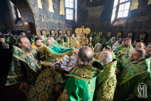 Eine Gruppe von Priestern in grünen Gewändern sitzt um einen Tisch in einer Kirche, mit verschiedenen Gegenständen auf dem Tisch und einer Wand mit Fenstern und Fotorahmen im Hintergrund, mit Text unten, der darauf hinweist, dass es ein Foto der Eparchie ist.