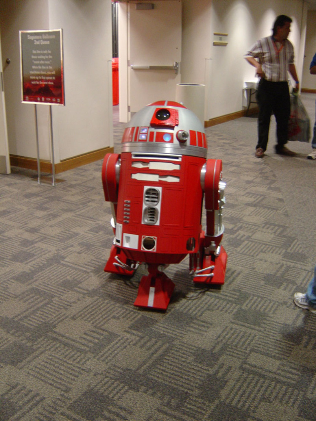 Ein Roboter auf dem Boden in der Vordergrund, mit einer Platte, Wand und Tür auf der linken Seite, drei Personen auf der rechten Seite und einem Tisch im Hintergrund.