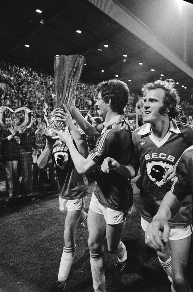 Eine Gruppe von Männern hält einen Pokal auf einem Fußballfeld, umgeben von einer Menge hinter einem Zaun, die ihren Sieg im FA-Cup-Finale feiert, dargestellt in Schwarz-Weiß.