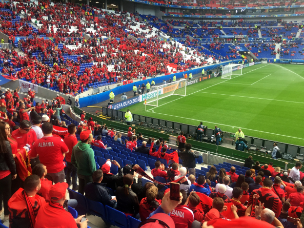 Ein volles Stadion mit sitzenden und stehenden Zuschauern, Bannern, einem Tor und Deckenleuchten, erfüllt von Erwartung auf ein Fußballspiel.