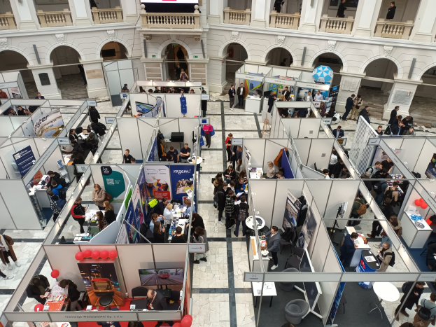 Große Gruppe von Besuchern auf einer internationalen Fachmesse mit Ständen, Bannern, Tischen, Stühlen und Ballons in einem Raum mit architektonischen Bögen und Säulen.