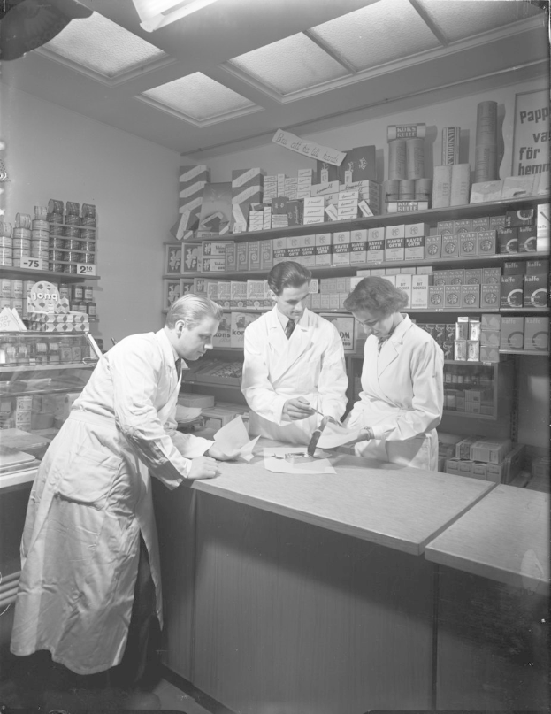 Drei Männer in weißen Kitteln an einer Apotheken-Theke mit Papieren und Gegenständen, mit Regalen voller Artikel im Hintergrund in einem Schwarz-Weiß-Bild.