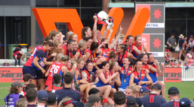 Gruppe von Frauen-AFL-Spielerinnen, die mit einem Pokal auf einem Rasenfeld feiern, tragen Sportuniformen und stehen vor einer Menge und einem Gebäude mit Glasfenstern.