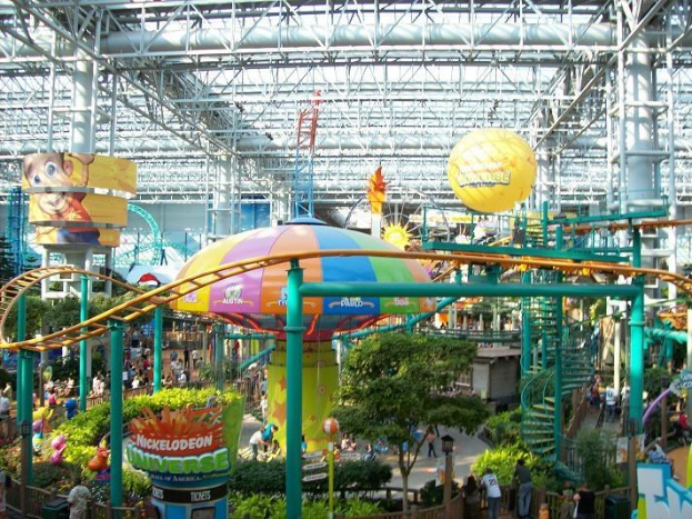 Großer Indoor-Wasserpark mit Achterbahn, umgeben von Menschen, Bäumen, Pflanzen und Deckenleuchten, die eine lebendige Atmosphäre schaffen.