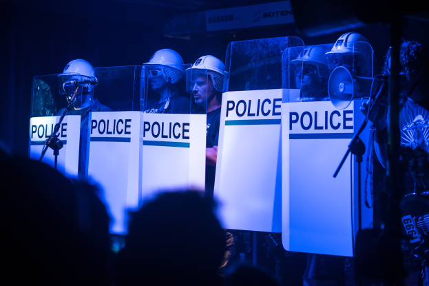 Eine Gruppe von Polizisten in Helmen steht vor einer Menge, hält Schilder mit Text, mit Mikrofonen und Ständern im Vordergrund und einem dunklen Hintergrund.