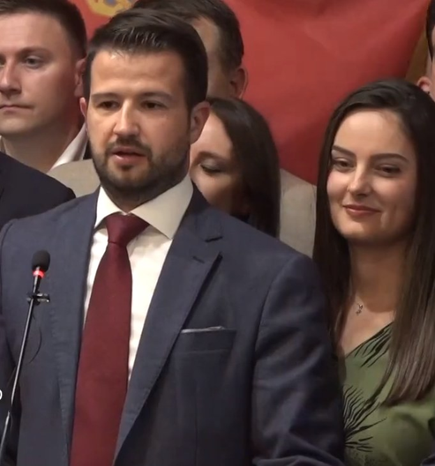 Ein Mann im Anzug spricht in ein Mikrofon an einem Rednerpult, mit einem Banner im Hintergrund und einer Menge, die likely auf ein Verbot der sozialen Medien durch die Regierung reagiert.