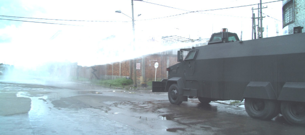 Ein schwarzer Lkw auf einer überfluteten Straße mit Rauch im Vordergrund, eine Wand rechts, ein Gebäude mit Pfählen und Drähten neben dem Lkw und ein Himmel darüber.