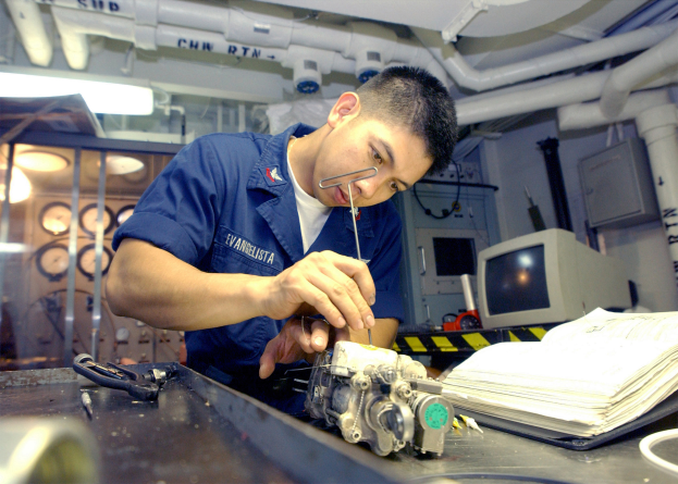 Ein Techniker in einem blauen Overall arbeitet an einer Maschine in einer Fabrik, mit Büchern, einer Maschine und anderen Gegenständen auf dem Tisch vor ihm und Lampen, Rohren und einer Wand im Hintergrund.