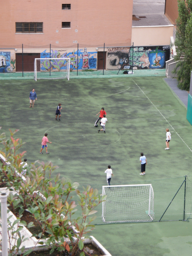Kinder beim Fußballspielen auf einem Feld mit Toren, im Hintergrund ein Gebäude mit Graffiti, links Pflanzen und rechts Bäume.