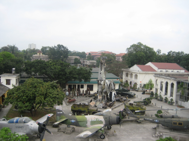 Drei Flugzeuge auf einer Landebahn mit Bäumen im Hintergrund, großen Häusern in der Nähe und einem klaren Himmel darüber.