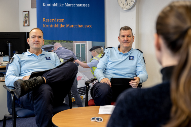 Zwei Polizeibeamte sitzen an einem Tisch mit einer Frau, die Dokumente durchsieht; eine Wanduhr und ein Banner, ein zweiter Tisch mit einem Monitor und Peripheriegeräten sowie Rahmenfotos sind im Hintergrund zu sehen.