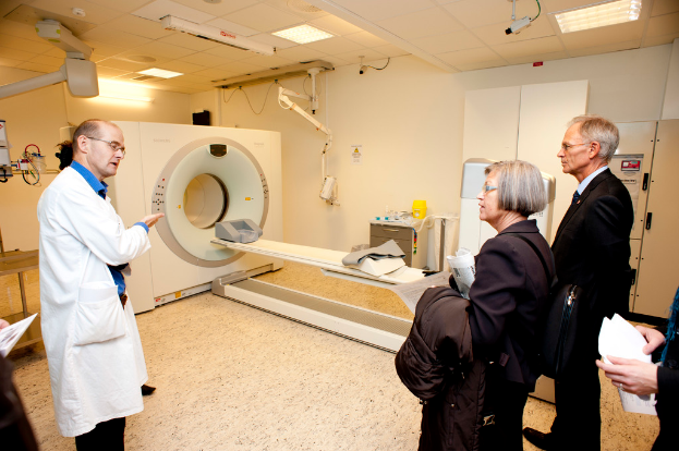 Ein Arzt in weißem Kittel erklärt eine CT-Scan-Maschine zwei Personen in dunklen Anzügen.