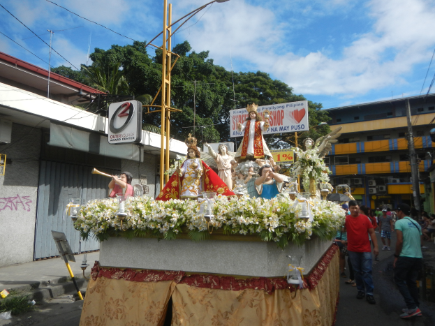 Ein Motivwagen mit Statuen, Blumen und Texttafeln, der durch eine Straße mit Laternenmasten, Kabeln, Gebäuden, Bäumen und einem bewölkten Himmel fährt.