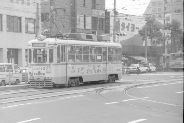 Ein Schwarz-Weiß-Foto einer belebten Stadtstraße mit einer Tram, Fahrzeugen, Radfahrern, Gebäuden mit Fenstern und Strommasten mit Drähten.