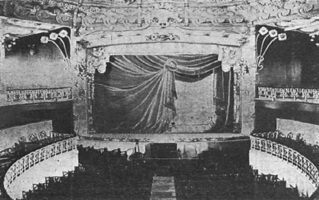 Schwarzes und weißes Foto des Auditoriums des Theatre Royal in London, das eine Bühne mit einem Vorhang, Geländern und kunstvollen Wanddesigns zeigt, mit einigen sitzenden Menschen und Text am unteren Rand.