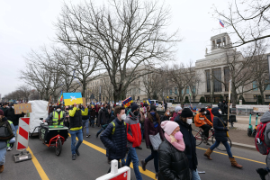 Eine große Gruppe von Menschen marschiert bei einer Demonstration auf einer Straße, einige halten Schilder und andere fahren Fahrräder, mit Bäumen und einem Gebäude im Hintergrund bei klarem blauem Himmel.
