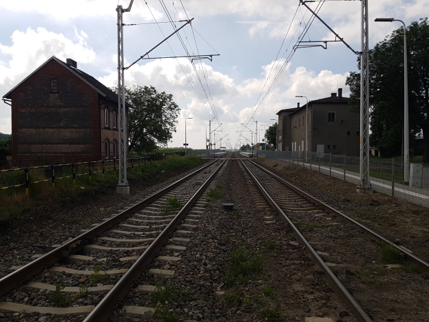 Eine Bahnstrecke mit Oberleitungen, umgeben von Gebäuden, Straßenmöbeln, Vegetation und einem bewölkten Himmel.