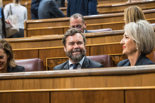 Eine Gruppe von Menschen, die in einem Gerichtssaal sitzen, mit einem Mann in einem Anzug und zwei Frauen in legerer Kleidung, die nebeneinander sitzen, mit Bänken im Hintergrund.