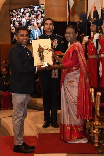 Ein Mann in einem schwarzen Blazer, weißem Hemd und grauen Hosen hält eine Trophäe, während er neben einer Frau in einem ashesfarbenen Sari auf einem roten Teppich steht, mit sitzenden und stehenden Gästen, Flaggen, Stühlen und einem wandmontierten Bildschirm im Hintergrund.