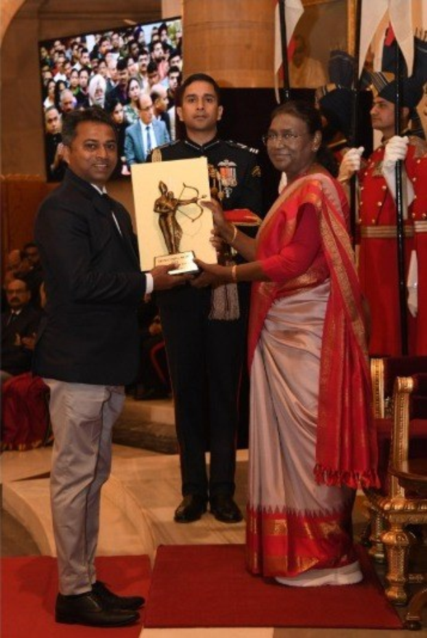 Ein Mann in einem schwarzen Blazer, weißem Hemd und grauen Hosen hält eine Trophäe, während er neben einer Frau in einem ashesfarbenen Sari auf einem roten Teppich steht, mit sitzenden und stehenden Gästen, Flaggen, Stühlen und einem wandmontierten Bildschirm im Hintergrund.