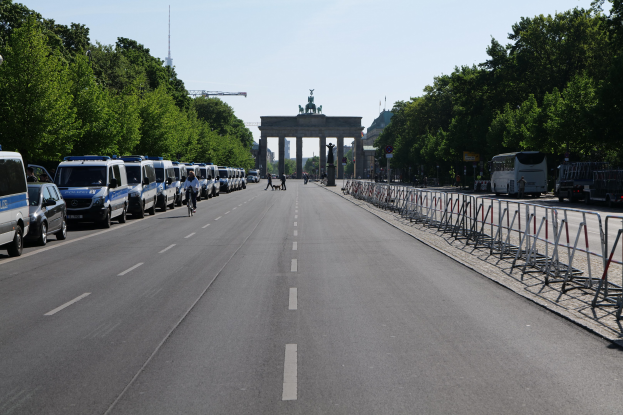 Eine Reihe von Polizeiwagen, die auf einer Straße vor dem Brandenburger Tor in Berlin, Deutschland, geparkt sind, mit Menschen auf Fahrrädern und in der Nähe stehenden Menschen, Absperrungen, Bäumen und einem Bogen mit Statuen im Hintergrund.