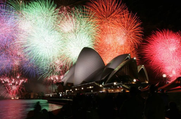 Das Sydney Opera House wird während der Neujahrsfeierlichkeiten von farbenfrohen Feuerwerken erhellt, während Menschen im Vordergrund die festliche Stimmung genießen.