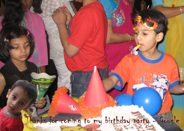 Kinder vor einem Tisch mit einem Kuchen, Luftballons und Mützen, mit Erwachsenen dahinter.