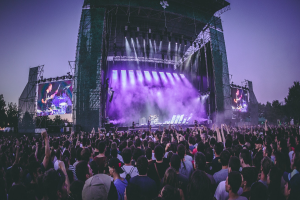 Eine große Menschenmenge bei einem Musikfestival schaut auf der beleuchteten Bühne mit Instrumenten und Bildschirmen, die vor einem Gebäude, Bäumen und einem blauen Himmel liegt.
