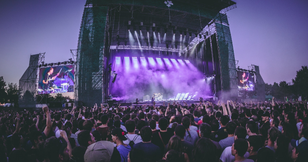 Eine große Menschenmenge bei einem Musikfestival schaut auf der beleuchteten Bühne mit Instrumenten und Bildschirmen, die vor einem Gebäude, Bäumen und einem blauen Himmel liegt.