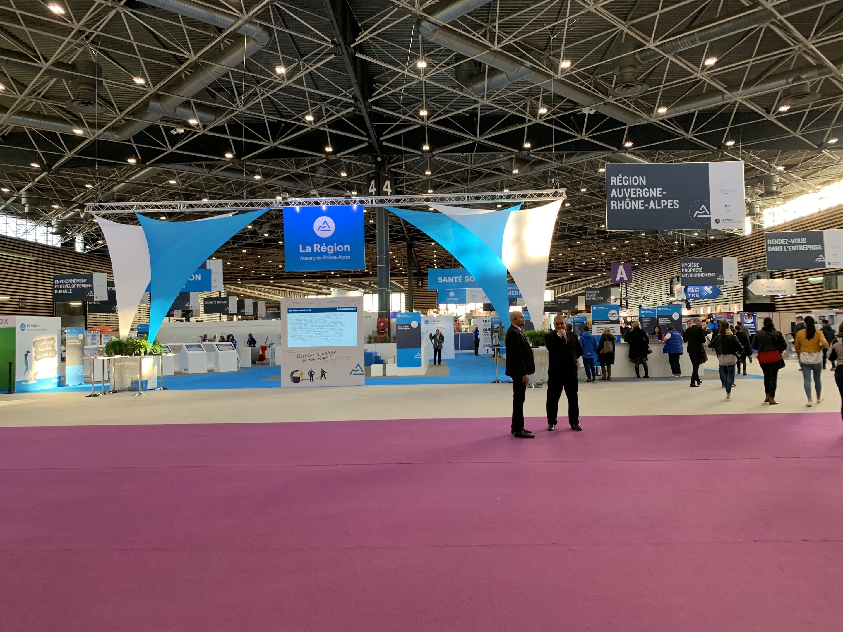 Ein großer Messestand auf der Pariser Internationalen Handelsmesse 2018 mit Menschen, die herumlaufen, von der Decke hängenden Bannern, Tischen mit Gegenständen und Deckenbeleuchtung.