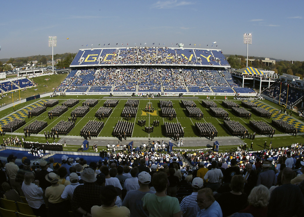 Eine große Menge schaut ein Fußballspiel in einem grünen Stadion unter einem klaren blauen Himmel, mit Laternen, Bäumen und Gebäuden im Hintergrund.