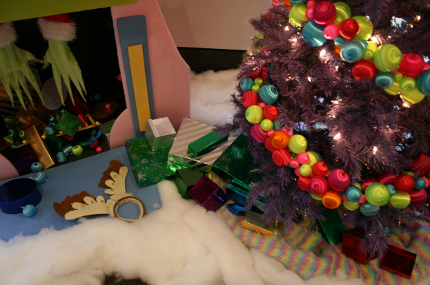 Drinnen steht rechts ein geschmÃ¼ckter Weihnachtsbaum mit Lichterkette und Schmuck, umgeben von Geschenkboxen und Deko-Artikeln, mit einer HÃ¼tte und Baumwolle im Hintergrund.