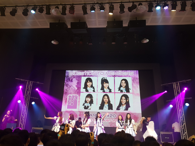 Eine Gruppe von Menschen, die auf einer hell erleuchteten Bühne performen, mit einer Menge darunter, die einen Sänger mit Mikrofon und einen Musiker mit Instrument zeigt, während ein großer Bildschirm Bilder der K-Pop-Gruppe BK48 anzeigt.