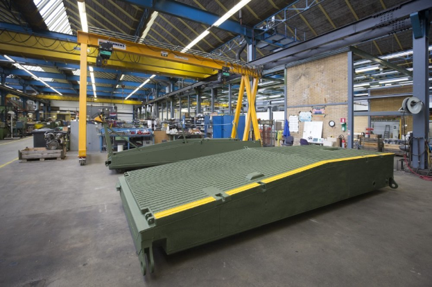 Ein große Lagerhalle mit zahlreichen grünen Behältern auf dem Boden, verschiedenen Maschinen, Polen, Lichtern und Stäben, mit einer Wand im Hintergrund, die eine Uhr und angeklebte Papiere zeigt.