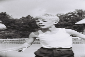 Ein junger Junge in Schwarz-Weiß spielt Tennis auf einem Court, schwingt einen Schläger in der Luft, mit einem Zaun, Bäumen und einem Haus im Hintergrund.