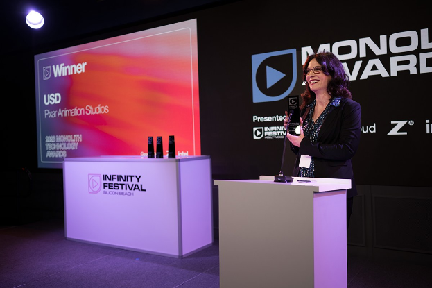 Frau in einem schwarzen Kleid, die auf einem Podium steht und einen Preis hält, mit einem Mikrofon und anderen Gegenständen auf dem Podium, vor einem Bildschirm und einer Werbetafel.