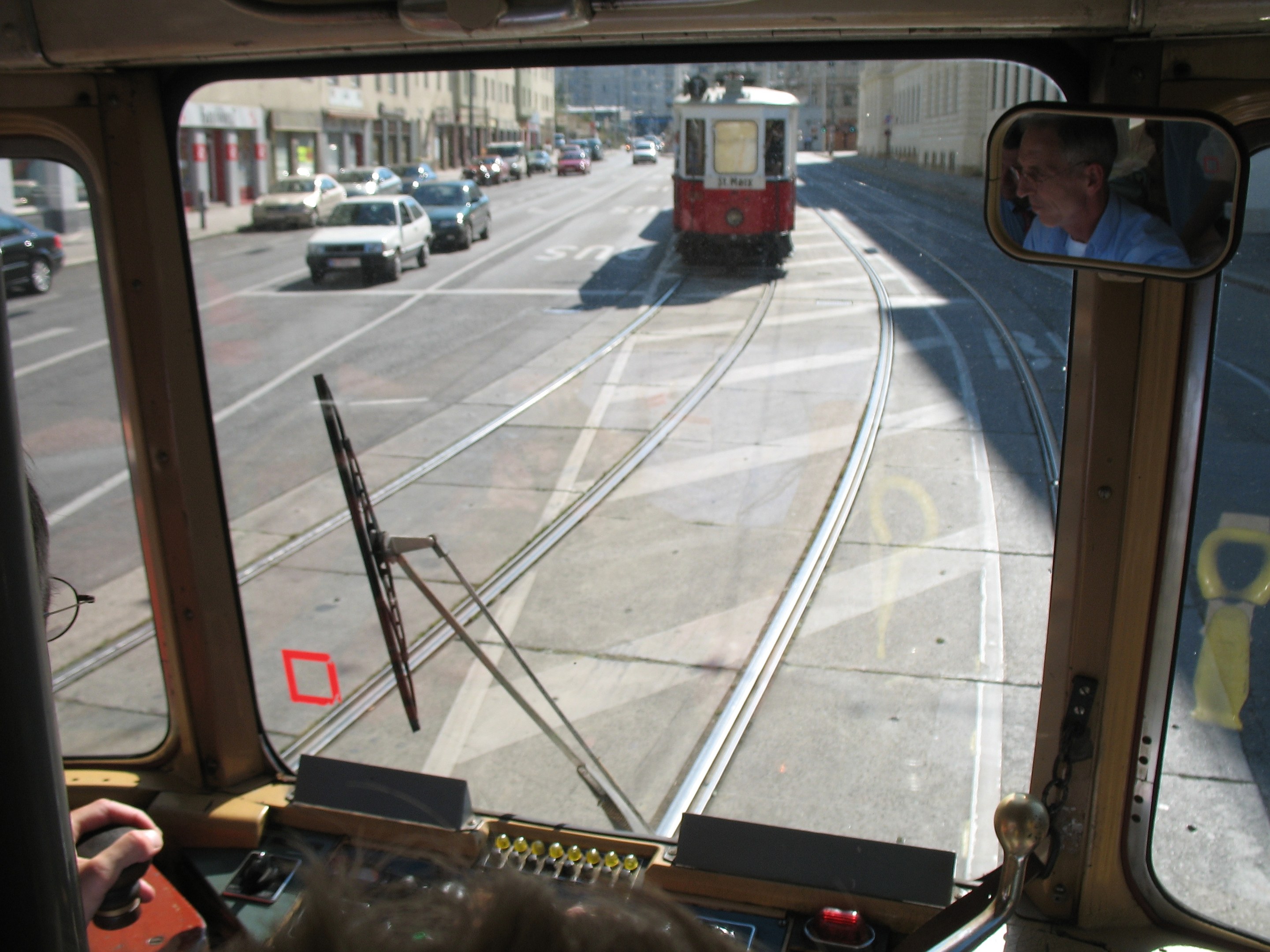 Ein Mann fährt eine Straßenbahn auf einer städtischen Straße mit anderen Fahrzeugen, Gebäuden und einem klaren blauen Himmel im Hintergrund.