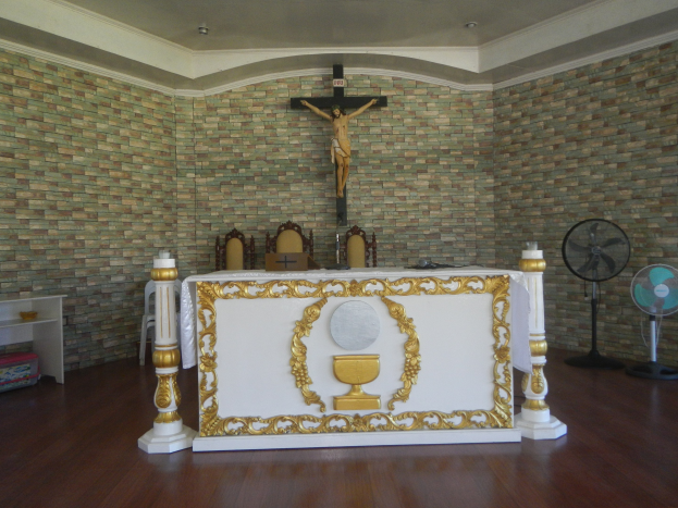 Altar in einer Kirche mit einem Kreuz an der Wand, einem Tisch mit Kerzen und anderen Gegenständen, um ihn herum angeordneten Stühlen, Gebläsen auf der rechten Seite und einem Gestell mit verschiedenen Gegenständen auf der linken Seite.