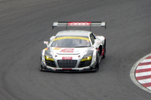 Ein leuchtend roter und weißer Audi R8 LMS GT3 Rennauto mit dem Audi-Logo auf der Seite, gefahren von einer Person in einem schwarzen Rennanzug und Helm, der um eine Rennstrecke fährt.