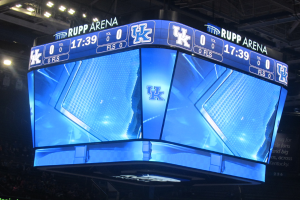 Ein großes Display in der Mitte eines Basketballfelds zeigt den Text "Kentucky Wildcats vs Florida State Seminoles", umgeben von Zuschauern auf den Tribünen und einer Decke mit Lampen darüber.