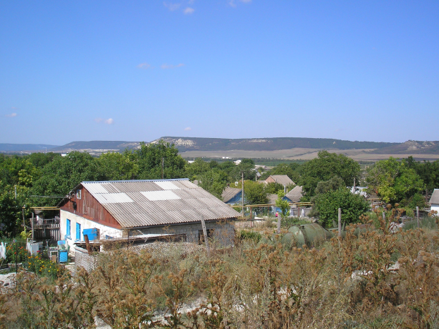 Kleines Dorf umgeben von Grün, mit Hügeln und einem klaren blauen Himmel im Hintergrund.