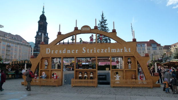 Ein geschäftiger Weihnachtsmarkt in Dresden, Deutschland, mit Menschen, die gehen, Taschen tragen und unter einem Bogen hindurchgehen, vor einem Hintergrund aus Bäumen, Gebäuden und einem bewölkten Himmel.