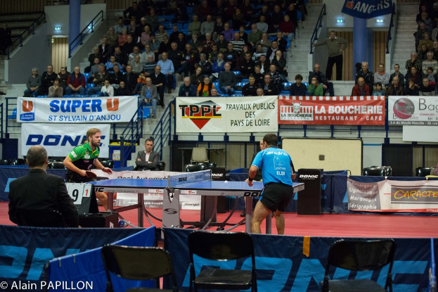 Zwei Männer spielen Tischtennis drinnen, mit mehreren Zuschauern, die stehen und sitzen, leeren Stühlen und Werbetafeln, mit Text in der unteren linken Ecke.