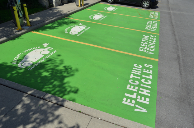 Eine Ladestation für Elektrofahrzeuge mit einem grünen Parkplatz, einem daneben geparkten Auto, gelben Pfosten und einer Wand auf der linken Seite, einem weiteren Auto und einem Gebäude im Hintergrund.