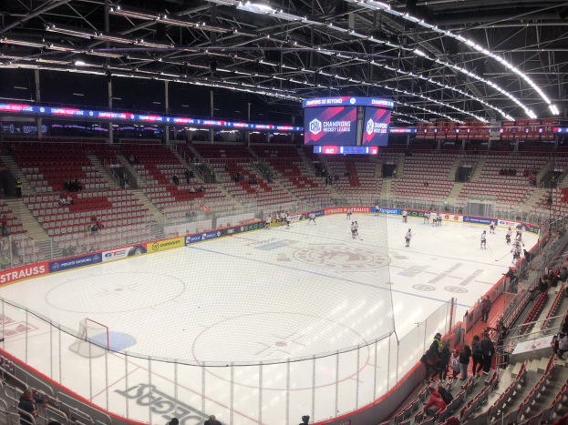 Ein Eishockeyspiel in einer großen Arena zwischen den St. Louis Blues und den Detroit Red Wings, mit Zuschauern auf den Sitzen und Stehplätzen in der Nähe des Spielfelds umgeben von einem Zaun, einem Bildschirm, Metallrahmen, Deckenleuchten und einem Dach.