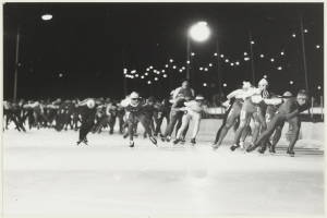 Eine Gruppe von Menschen, die nachts auf einer Eisbahn Schlittschuh laufen, im Hintergrund von Straßenlaternen beleuchtet, auf einem Schwarz-Weiß-Foto.